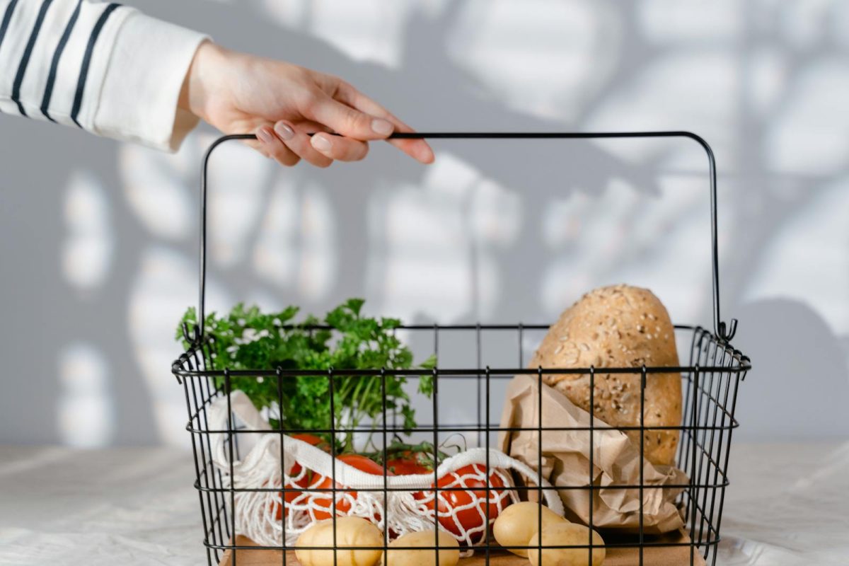 A hand holding a metal basket filled with eco-friendly groceries like bread, vegetables, and parsley.